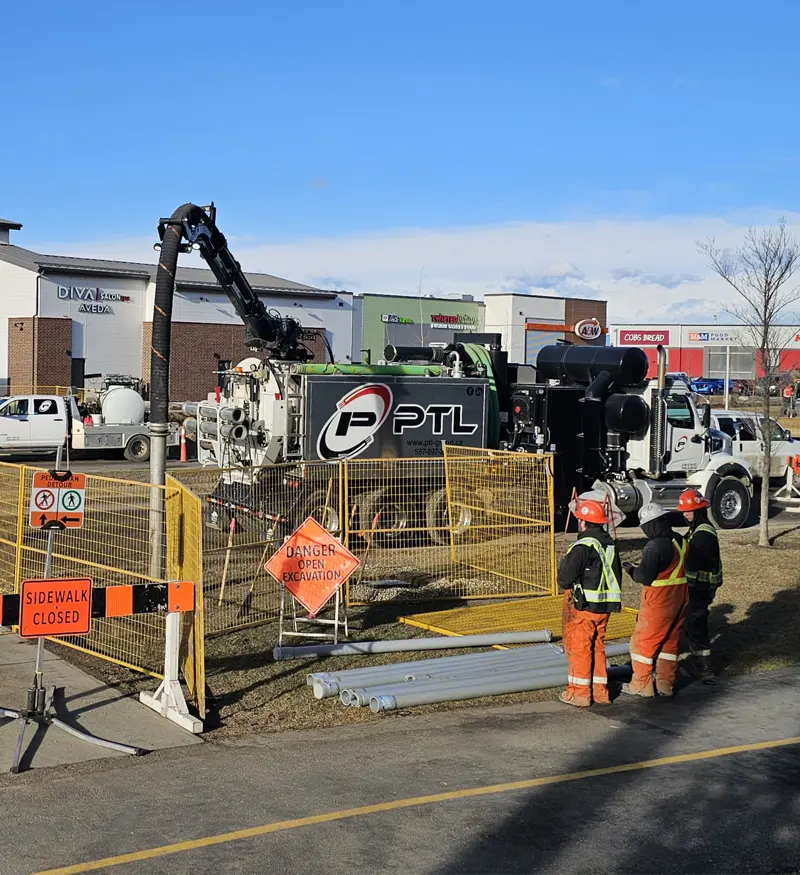 comprehensive-civil-utility-construction White and black hydrovac truck with its vacuum boom deployed next to two utility workers and a yellow safety fence at a sidewalk excavation site.