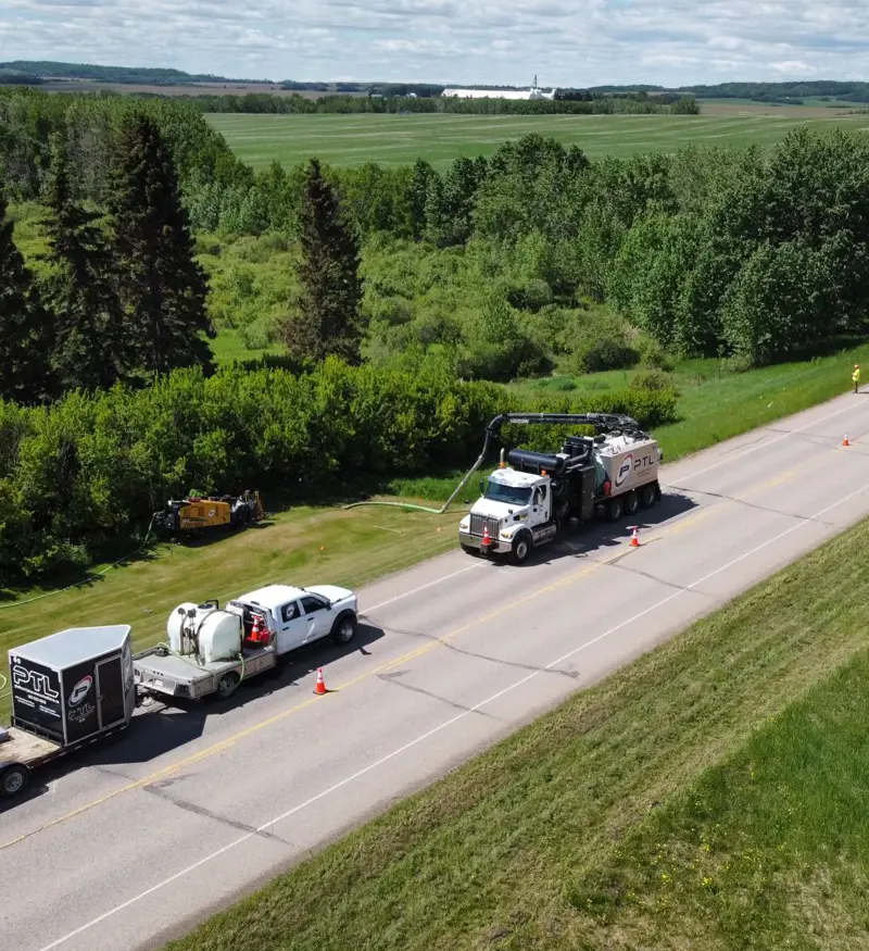 environmental-fluid-containment-site-restoration Aerial view of a white fluid management support truck and a hydrovac vehicle staged on a roadway shoulder with vacuum lines extended into a grassy ditch.