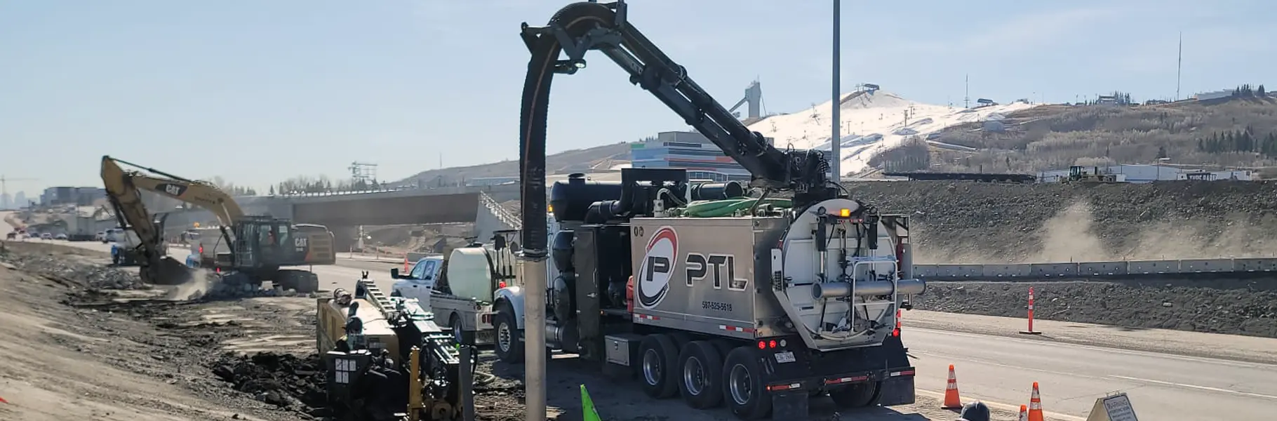 White tri-axle hydrovac truck with its long black vacuum boom arm extended high into the air on a roadside job site with a snowy mountain slope in the background.