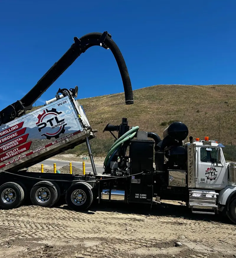 hydrovac-excavation-utility-exposure White tri-axle hydrovac truck with its black vacuum boom arm extended upward on a dirt utility job site next to a dry, grassy hillside.