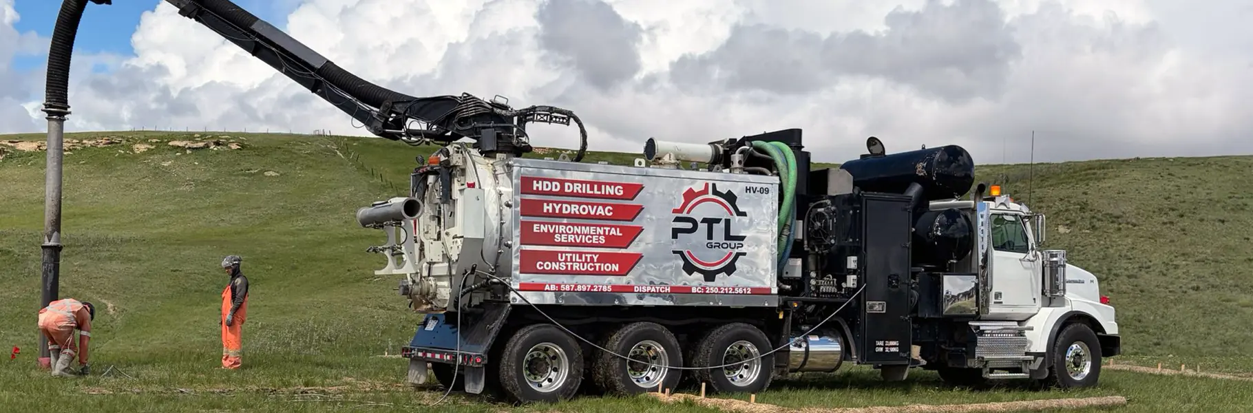 White tri-axle hydrovac truck with its vacuum boom extended, parked on a grassy slope next to utility workers in orange safety vests.