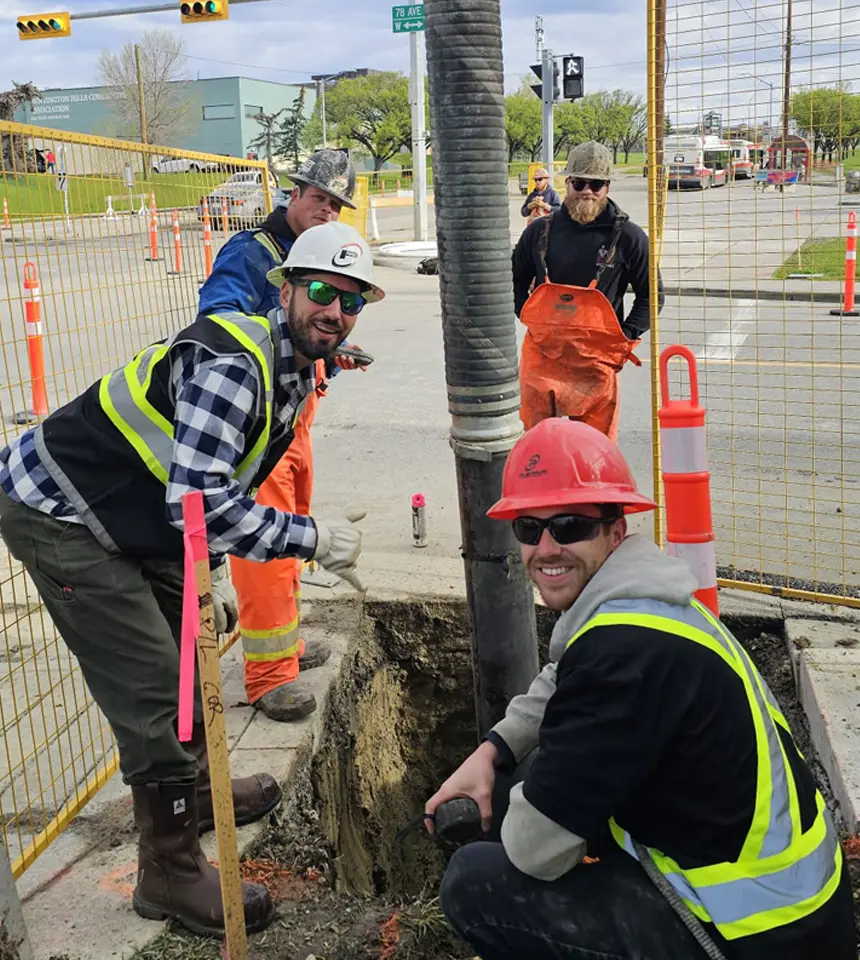 Three civil utility construction workers wearing hard hats and safety vests collaborating around an open sidewalk excavation pit on a city street.