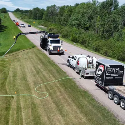 White environmental fluid management truck and trailer setup parked on a roadway shoulder with a long green suction hose extending across a grass ditch.