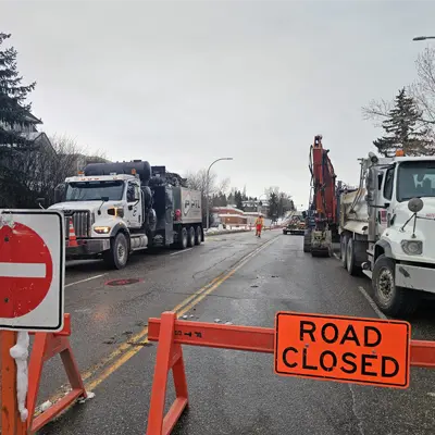 Active civil utility construction site along a municipal roadway corridor with traffic control barricades and underground excavation machinery.