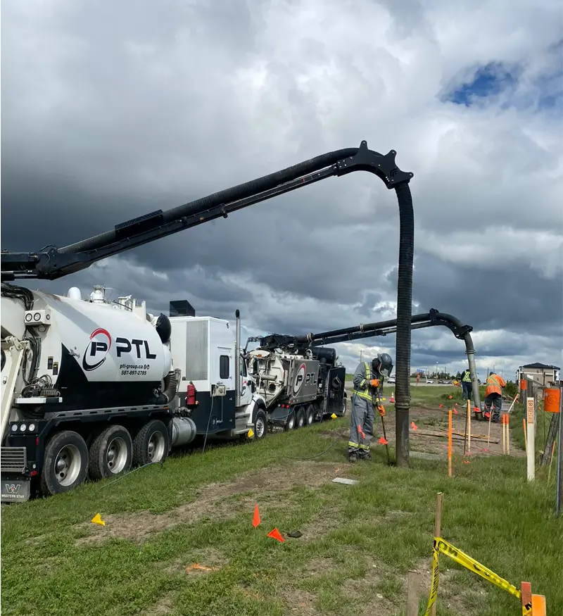 hydrovac-truck-boom-operation-utility-daylighting Silver high-capacity hydrovac truck with fully extended vacuum boom being operated by a crew member over a tank on a utility construction site.